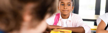 horizontal crop of african american schoolgirl sitting in school dining room near lunch boxの写真素材