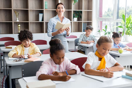 teacher holding book and looking at camera while multicultural pupils writing dictation in classroomの写真素材