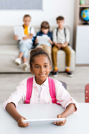 Selective focus of african american schoolgirl with backpack holding digital tablet at desk near...