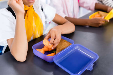 cropped view of schoolgirl taking fresh carrot from lunch box near african american classmateの写真素材