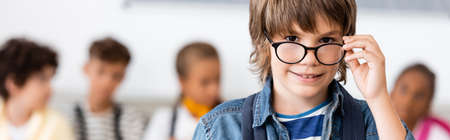Panoramic concept of schoolboy holding eyeglasses and looking at camera in school