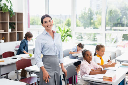 teacher looking at camera while standing in classroom near multicultural pupils