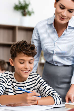 Selective focus of teacher standing near pupil holding pen near notebook at desk in classroomの写真素材