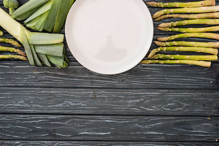 top view of fresh green leek, asparagus and empty plate on wooden surfaceの写真素材