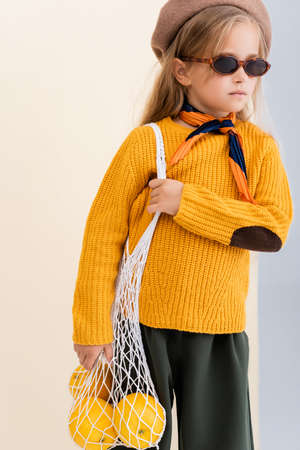 fashionable blonde girl in autumn outfit and sunglasses posing with grapefruits in string bag on...