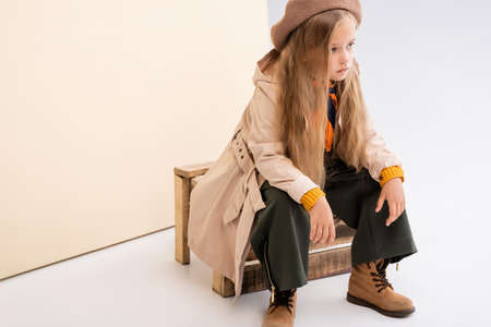 fashionable blonde girl in autumn outfit sitting on wooden box on beige and white backgroundの写真素材