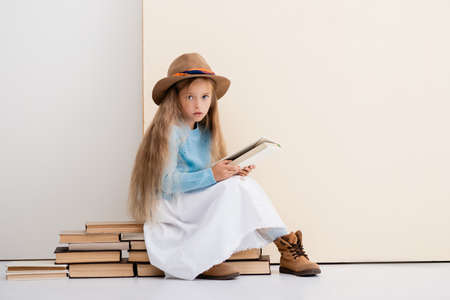 fashionable blonde girl in brown hat and boots, white skirt and blue sweater sitting on vintage books and reading near beige wallの写真素材