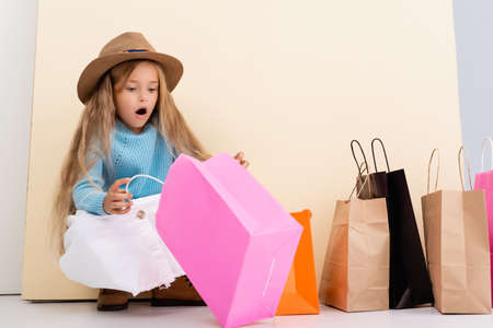 shocked fashionable blonde girl in brown hat and boots, white skirt and blue sweater looking inside colorful shopping bag near beige wallの写真素材