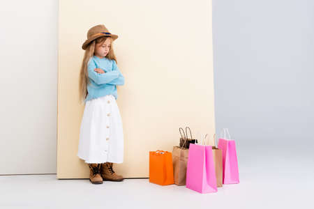 sad fashionable blonde girl in brown hat and boots, white skirt and blue sweater near colorful shopping bags and beige wallの写真素材