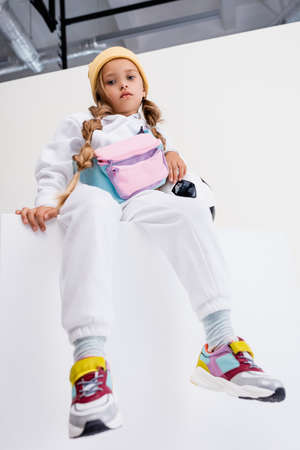 low angle view of blonde girl in sportswear sitting on cube with soccer ballの写真素材