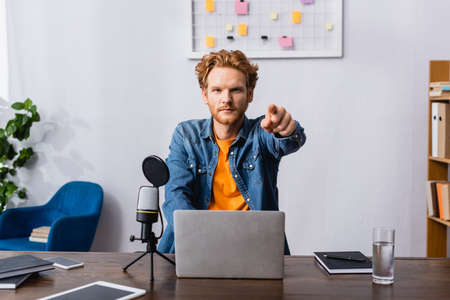 serious radio host in denim shirt pointing with finger at camera while sitting at workplaceの写真素材