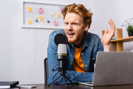 tense broadcaster in denim shirt gesturing while speaking in microphone near laptop