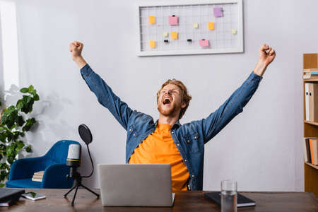 excited radio host showing triumph gesture while screaming near microphone and laptop
