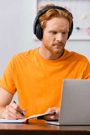 concentrated freelancer in wireless headphones writing in notebook while looking at laptopの写真素材