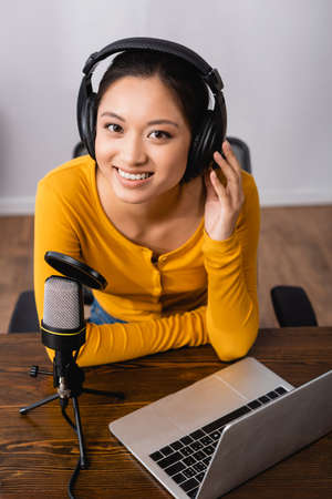 joyful asian announcer touching wireless headphones while sitting near microphone and laptop in studioの写真素材