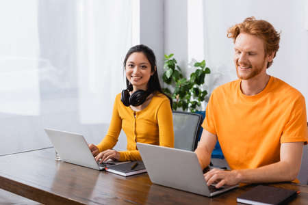 brunette asian freelancer with wireless headphones on neck looking at camera while using laptop near...