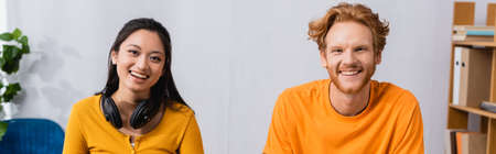 horizontal image of brunette asian woman with wireless headphones on neck, and redhead man looking at camera togetherの写真素材