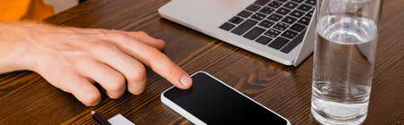 cropped view of teleworker touching smartphone with blank screen near laptop and glass of water,...