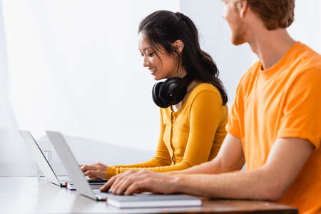 selective focus of freelancer using laptop near asian woman with wireless headphones on neckの写真素材