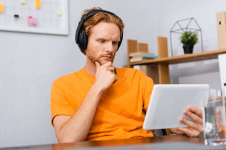 selective focus of thoughtful student in wireless headphones touching chin while using digital tabletの写真素材