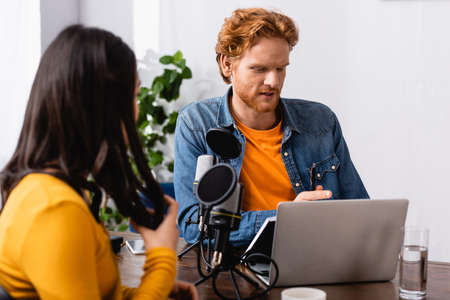 brunette woman touching hair while sitting near microphone and redhead radio host in studioの写真素材