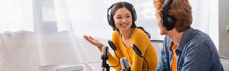 horizontal image of asian announcer in wireless headphones gesturing while talking to colleague in studioの写真素材