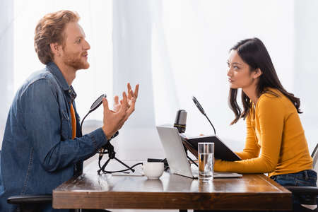 side view of redhead man gesturing while talking to young asian radio host in studioの写真素材