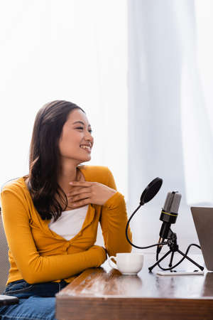 excited asian broadcaster touching chest while sitting near microphone at workplaceの写真素材
