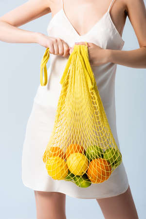 cropped view of woman in silk dress holding string bag with fruits on white, ecology conceptの写真素材