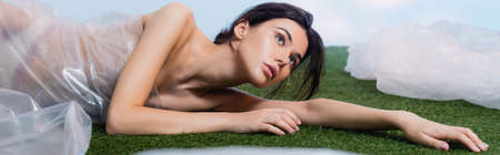 horizontal crop of brunette woman wrapped in polyethylene lying on grass, ecology conceptの写真素材