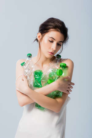 brunette woman in silk dress holding plastic bottles and looking down isolated on gray, ecology conceptの写真素材