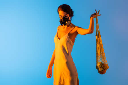young brunette woman in gas mask and silk dress holding reusable string bag with fruits on blueの写真素材