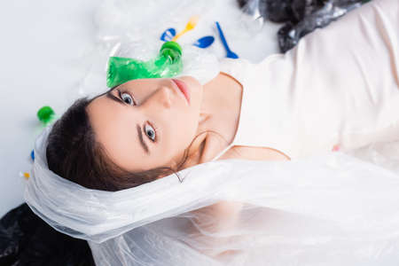 top view of woman with plastic bag on head sitting near empty bottles and spoons on white, ecology conceptの写真素材