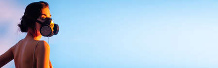 panoramic crop of brunette woman in gas mask looking at camera on blue