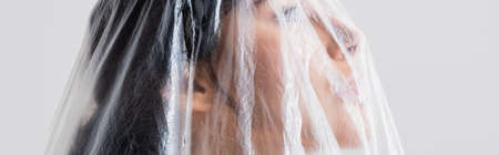 panoramic crop of young brunette woman looking through polyethylene isolated on gray, ecology conceptの写真素材
