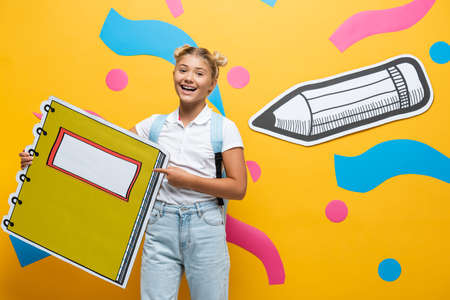 excited pupil pointing with finger at notebook maquette on yellow background with paper cut pencil...