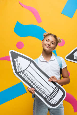 Schoolkid looking at camera while holding paper pencil on yellow background