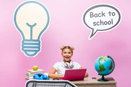 Child in headphones sitting near laptop, globe and paper artwork on pink backgroundの写真素材