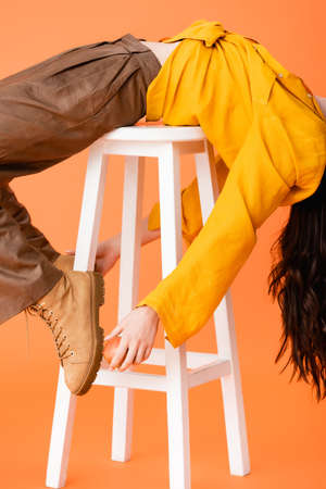 cropped view of trendy woman in autumn outfit lying on white stool and touching boots on orangeの写真素材