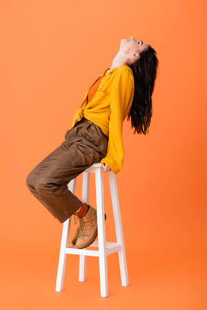 trendy woman in autumn outfit sitting on white stool and looking up on orange
