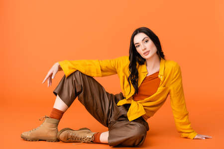 fashionable young woman in autumn outfit and boots looking at camera while sitting on orangeの写真素材