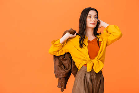 trendy woman in glasses and autumn outfit holding leather jacket while standing isolated on orangeの写真素材