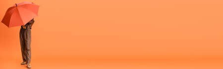 panoramic shot of fashionable woman in autumn outfit and boots covering face with umbrella on orangeの写真素材
