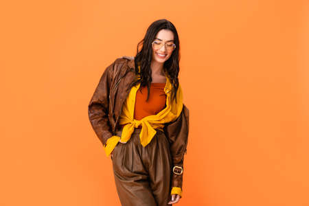 young woman in autumn outfit standing with hand in pocket and looking down isolated on orangeの写真素材