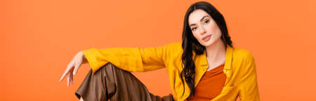 horizontal image of brunette young woman in autumn outfit looking at camera isolated on orangeの写真素材