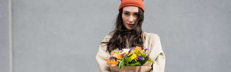 panoramic shot of stylish woman in beanie hat and trench coat holding flowers near gray wallの写真素材