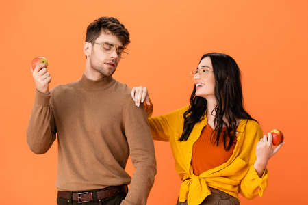 stylish couple in glasses and autumn outfit holding apples while looking at each other isolated on...