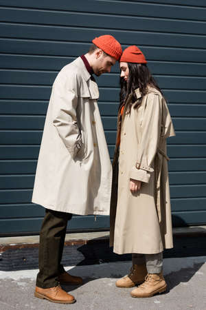 full length of stylish couple in trench coats and hats leaning on each other outsideの写真素材