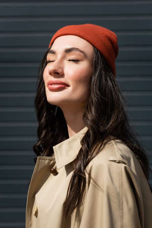 sunshine on stylish woman with closed eyes in trench coat and beanie hat