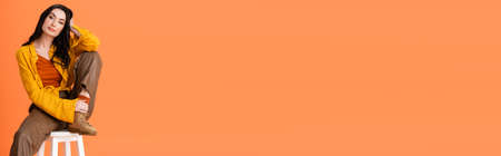 horizontal image of young woman in autumn outfit sitting on white stool isolated on orangeの写真素材